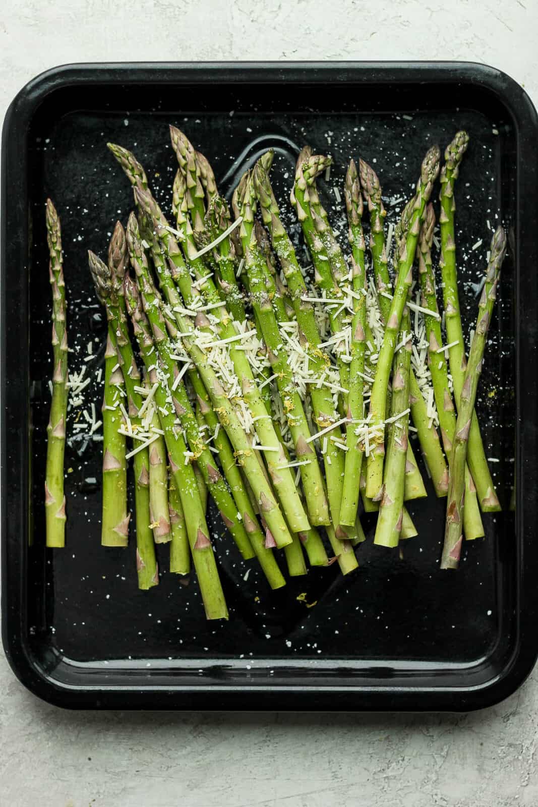 Trimmed asparagus on a black baking sheet sprinkled with kosher salt and parmesan.