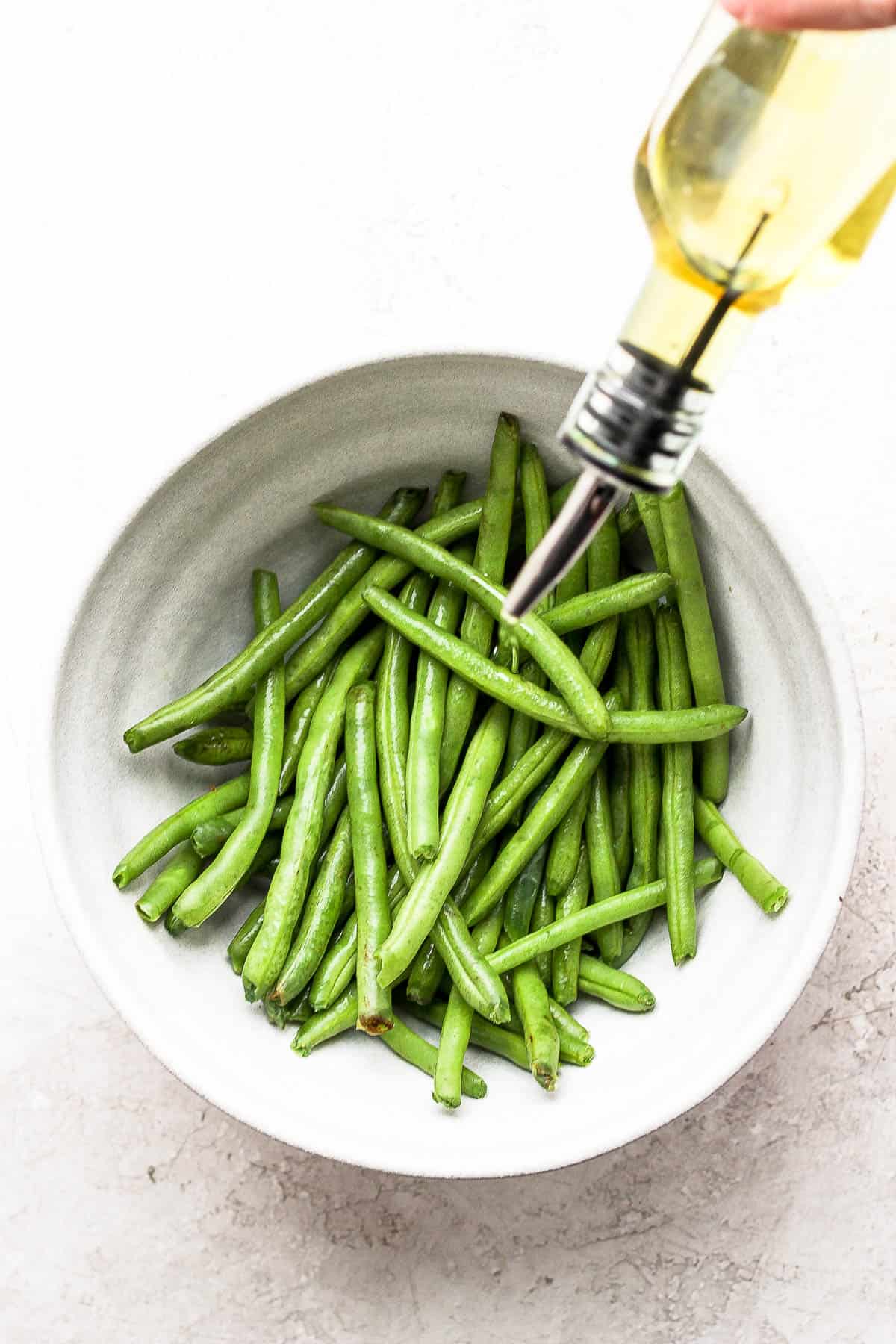 Plain green beans in a white bowl being drizzled with olive oil.