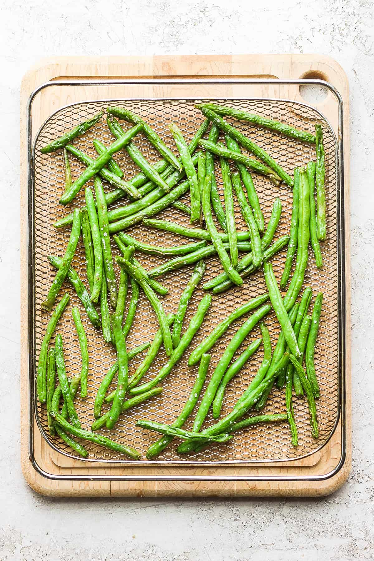 Air fried green beans in an air fryer basket set on a cutting board. 