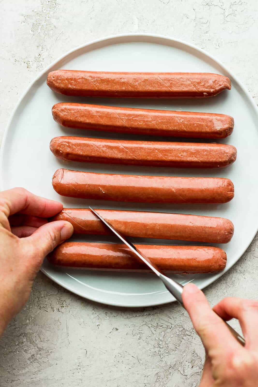 A paring knife scoring one of six hot dogs on a plate.
