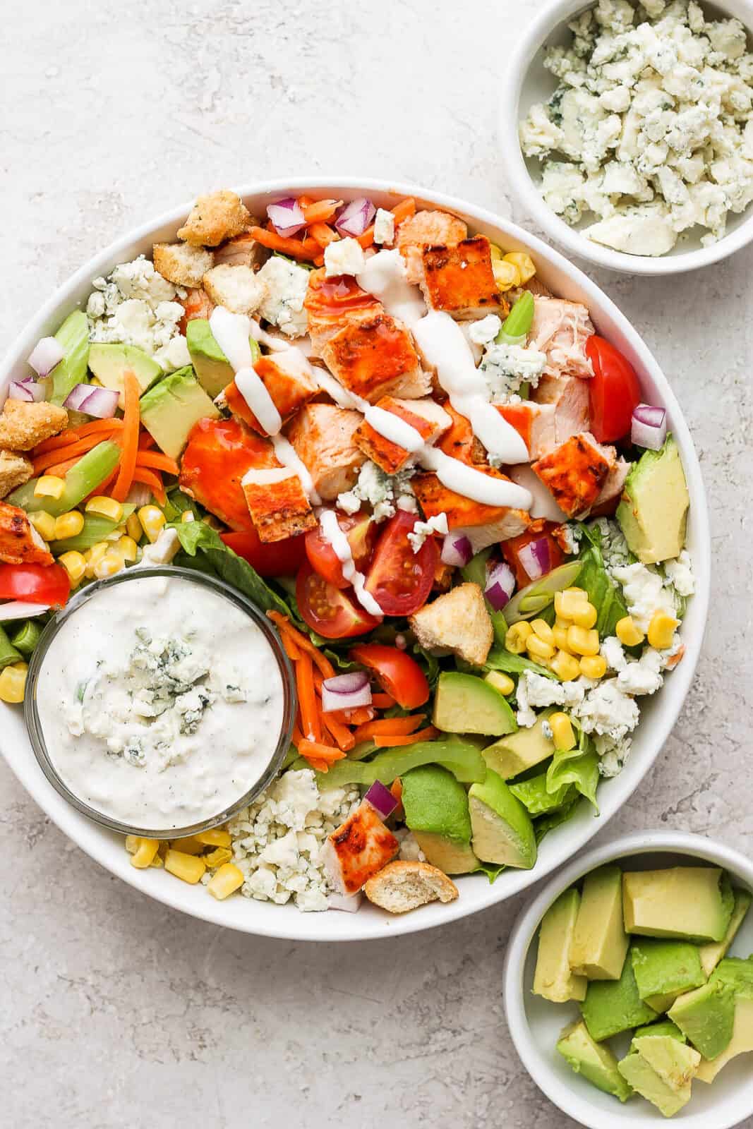 A buffalo chicken salad with small bowls of dressing, avocado, and blue cheese crumbles.