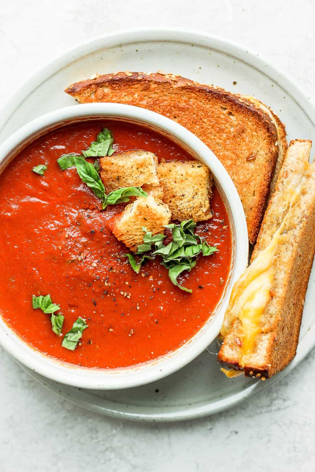 A bowl of creamy tomato basil soup with homemade croutons on top and an air fryer grilled cheese on the side.