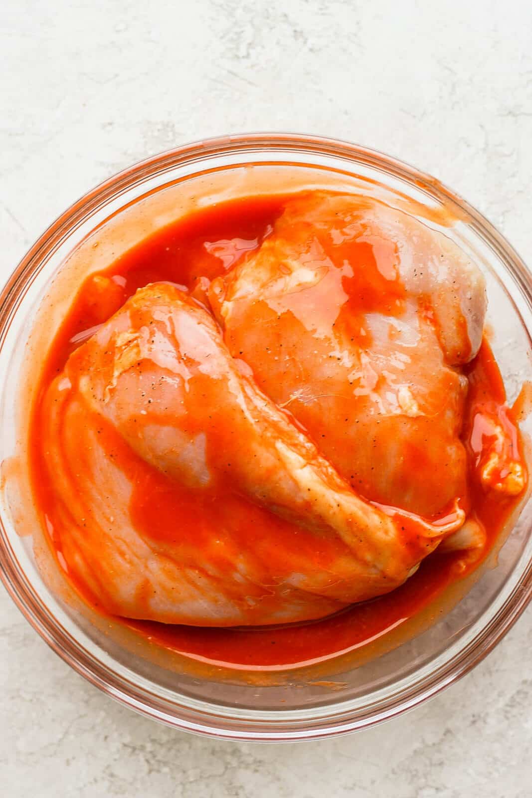 Chicken breasts marinating in a bowl with buffalo sauce.