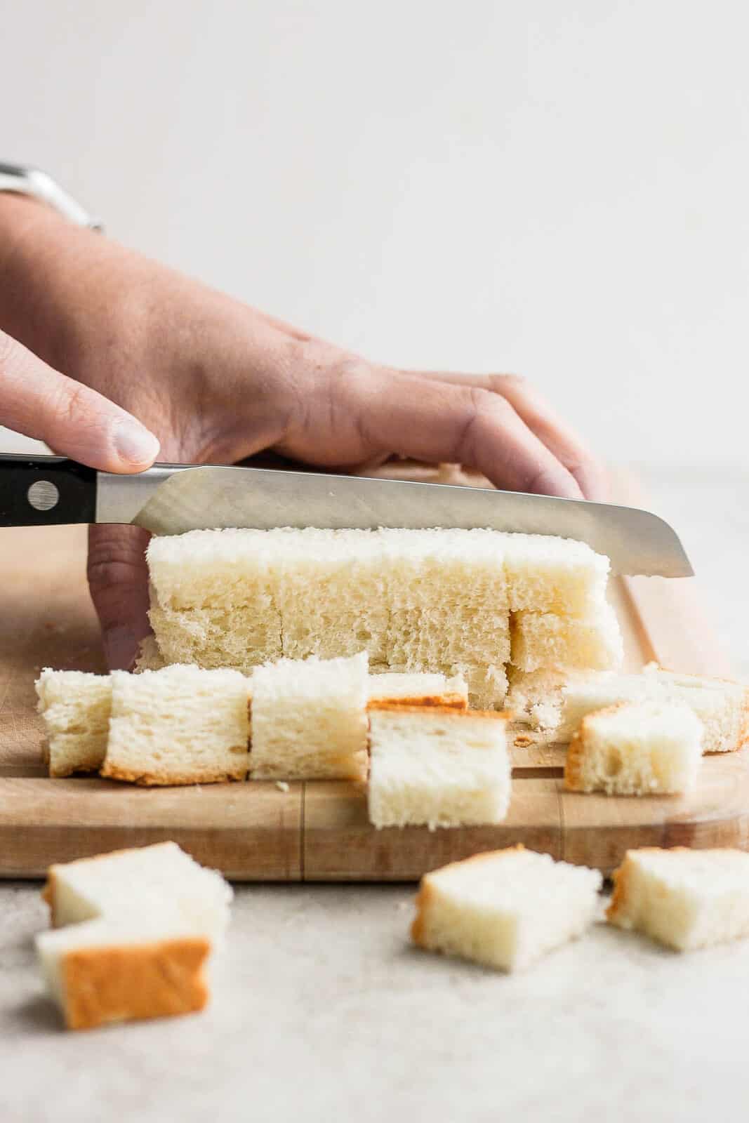 Bread being cubed using a serrated knife on a cutting board.