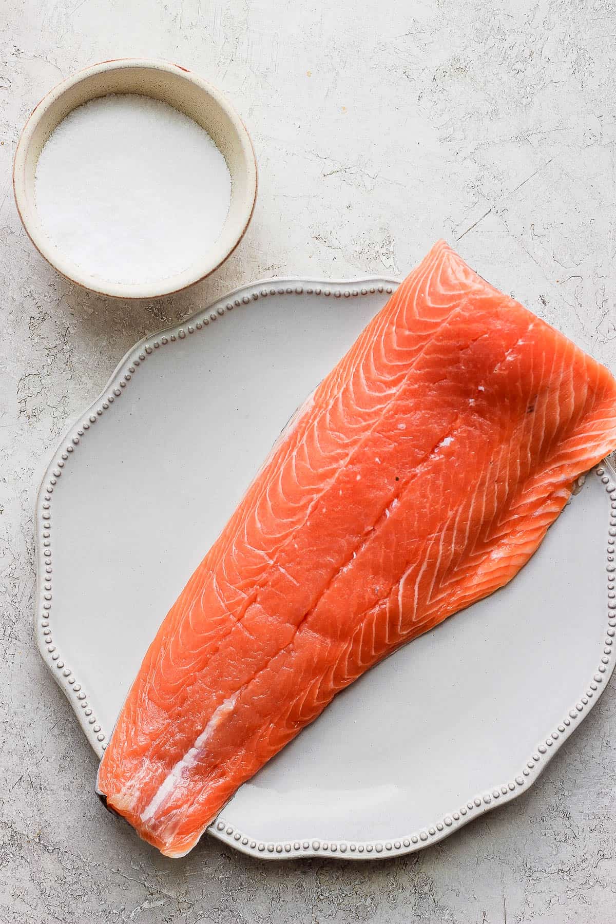 A bowl of kosher salt and a fillet of salmon on a plate. 