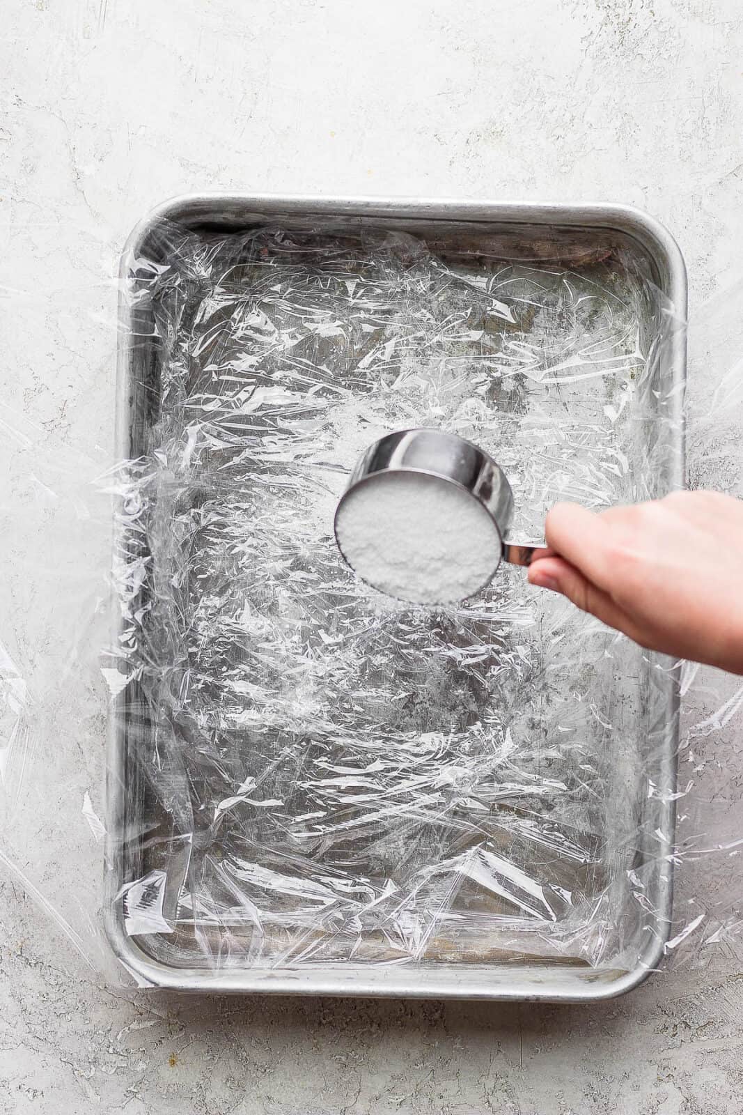 Someone pouring salt into a plastic wrap-lined baking dish. 