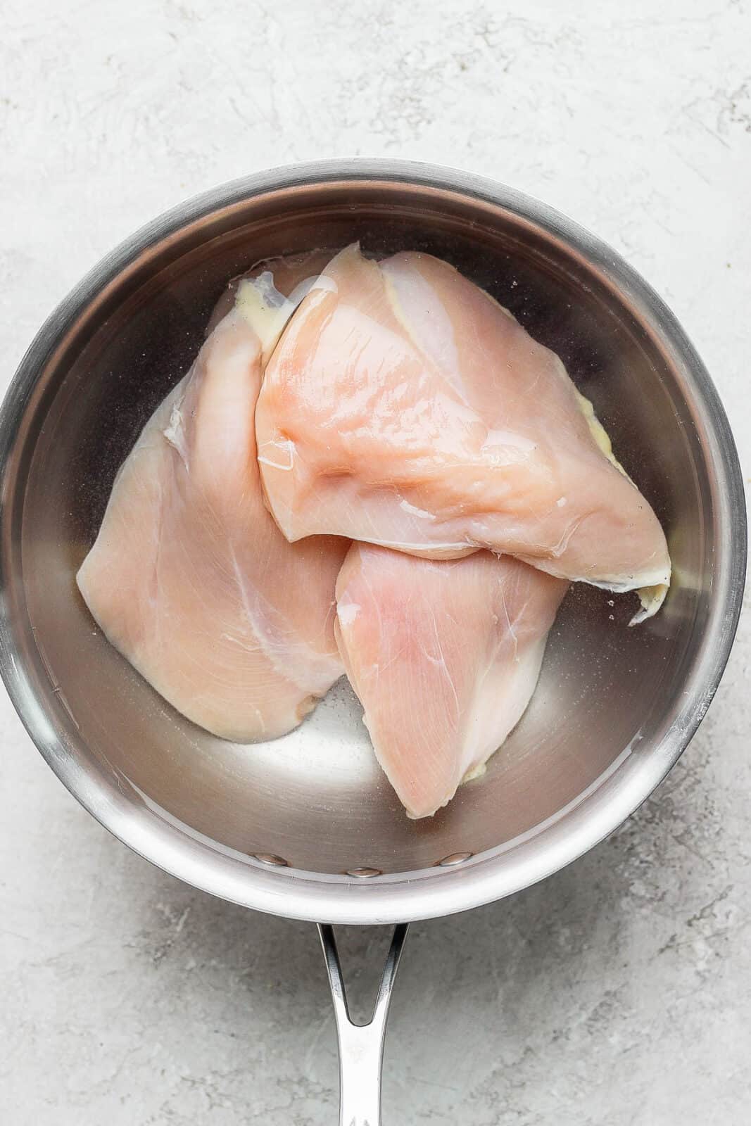 A large pot with water with three chicken breast inside.