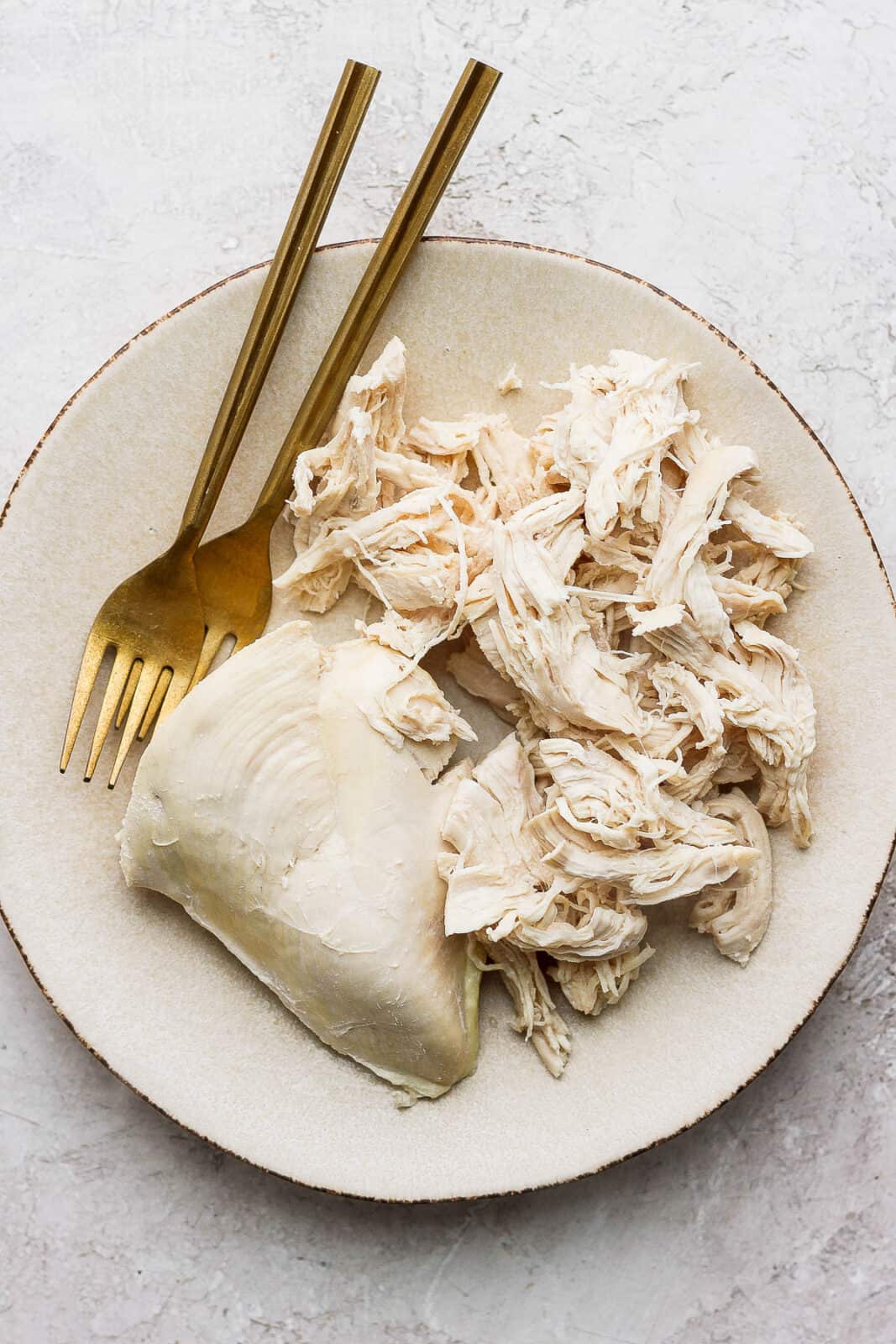 A white plate with two gold forks for shredding, shredded poached chicken, and part of a whole chicken breast.