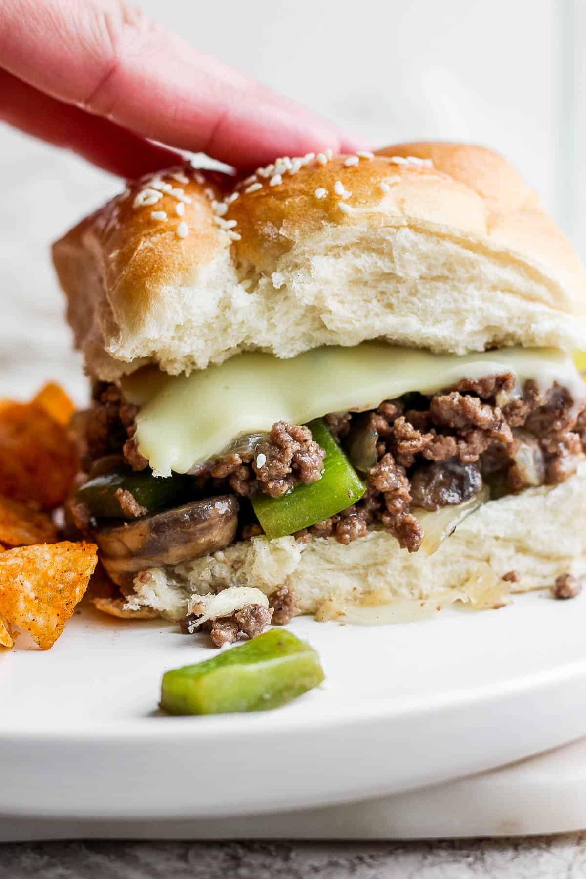 A hand touching the top bun of a philly cheesesteak sloppy joe.