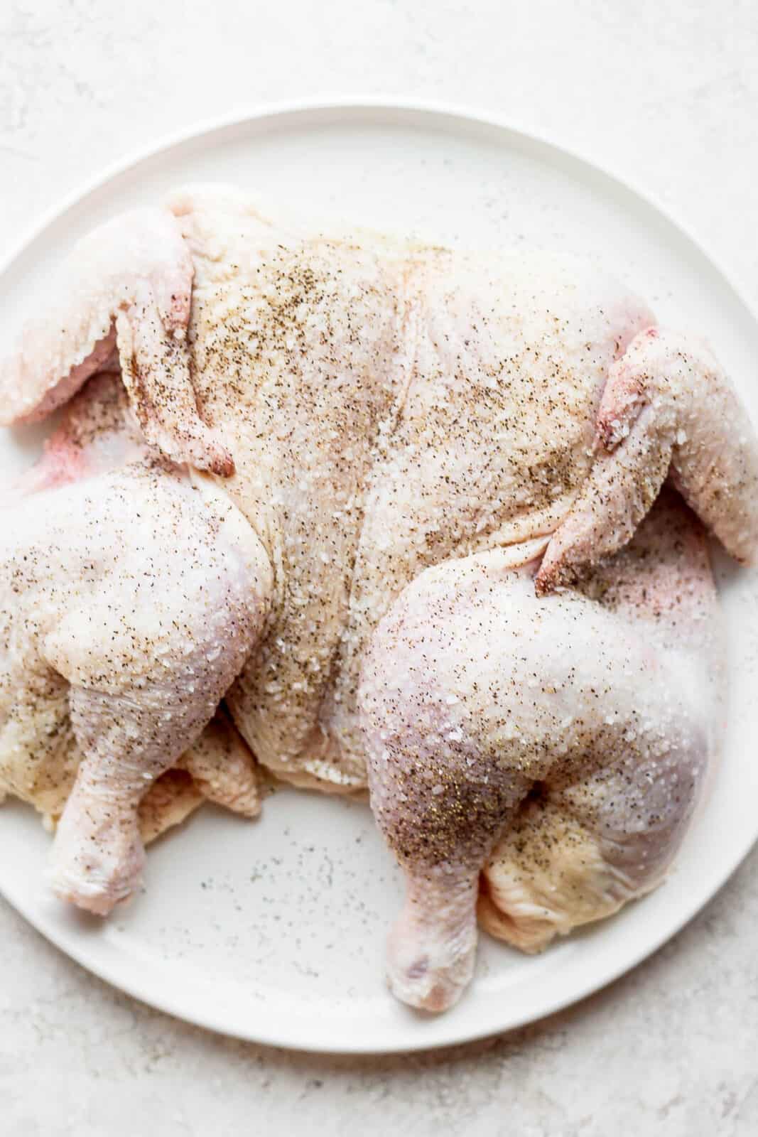 A raw spatchcocked chicken, seasoned, on a plate.
