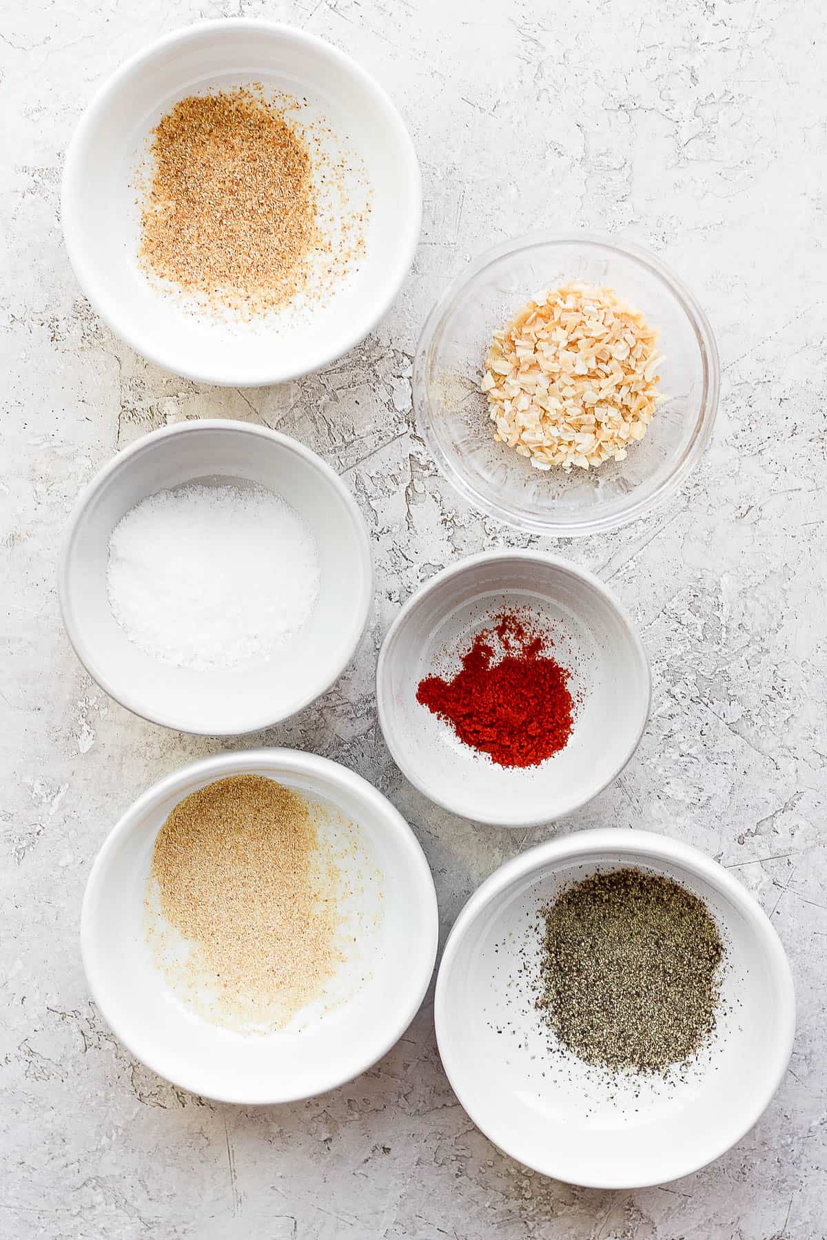 Individual small bowls filled with each seasoning ingredient; kosher salt, ground pepper, paprika, onion powder, garlic powder, and dried onion. 