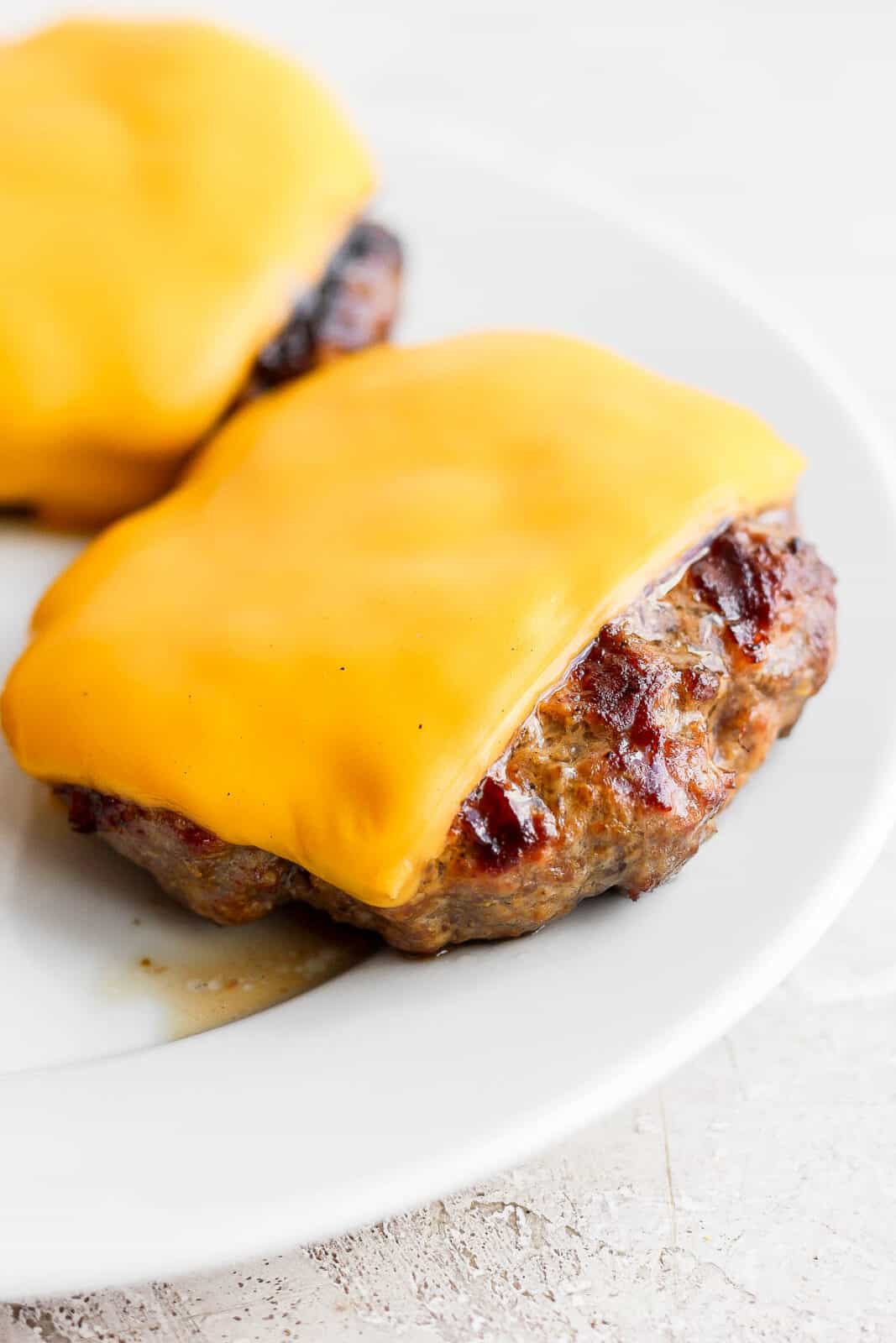 Two cooked burger patties topped with cheddar cheese on a white plate. 