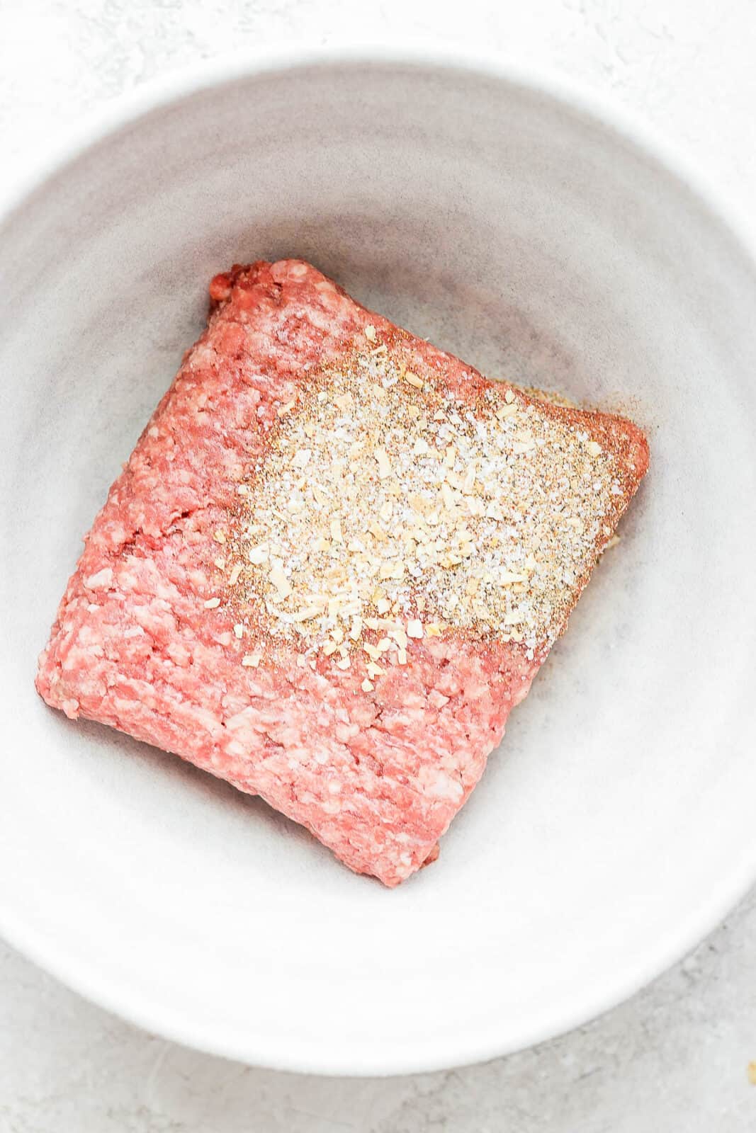 Burger seasoning on top of ground meat in a white bowl. 