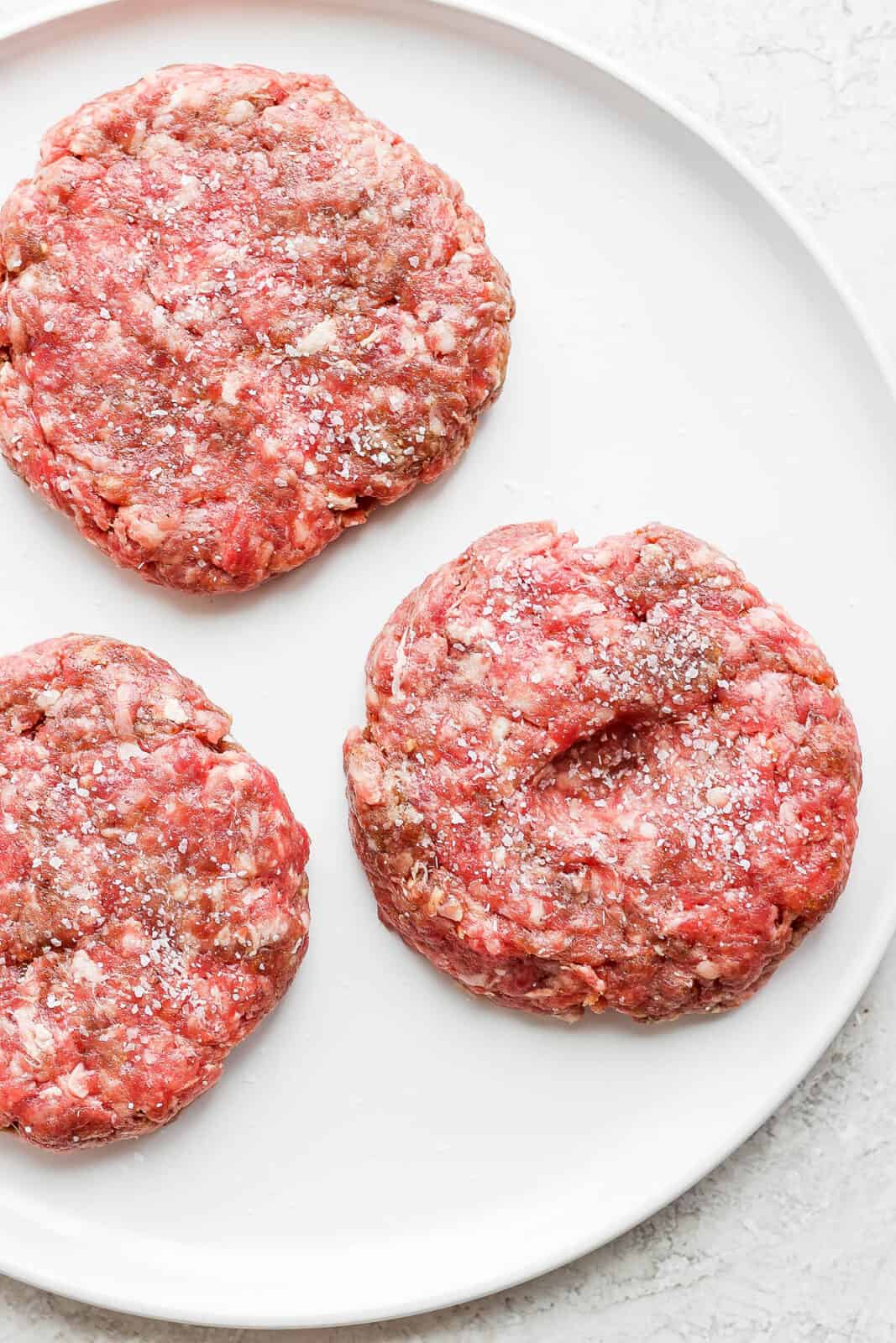 A plate with 3 burger patties seasoned with salt and pepper. 