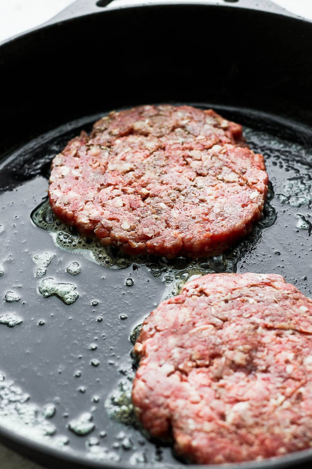 A close up of two patties cooking in a cast iron skillet. 