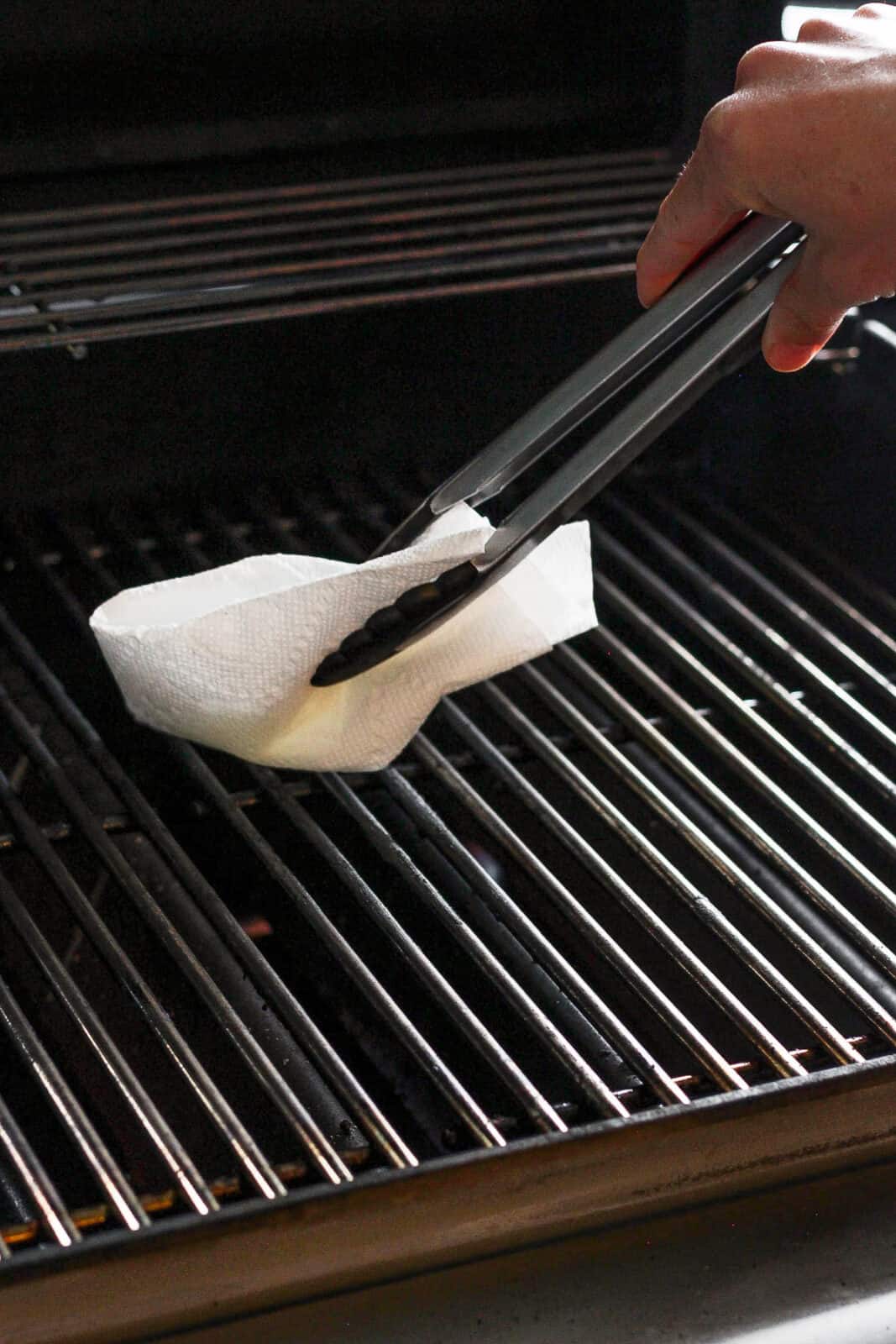 Hot grill grates being rubbed down with a paper towel that has olive oil on it.
