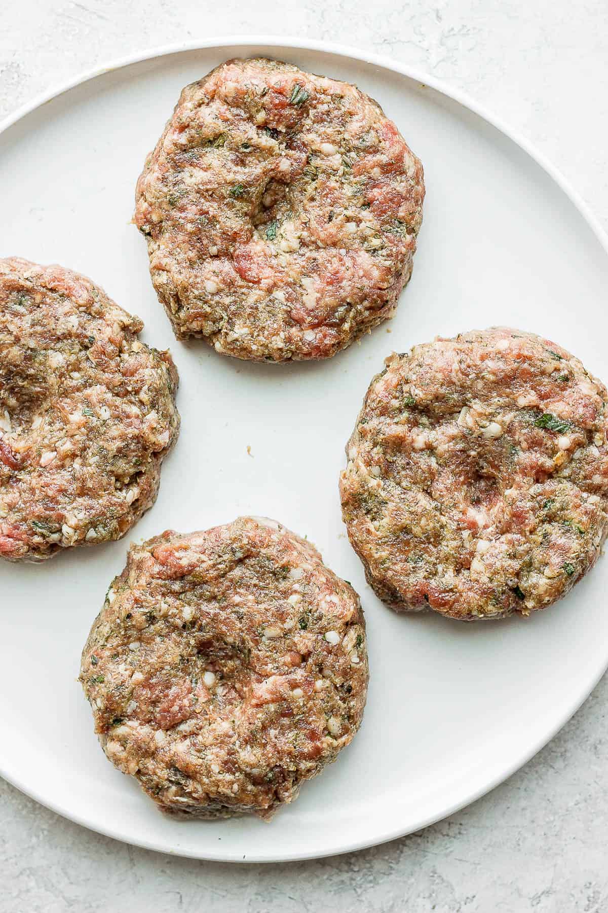 Four lamb burger patties on a plate with thumb indents in them.