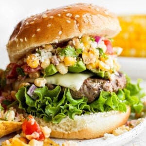 A burger on a bun with lettuce and mexican street corn salad.