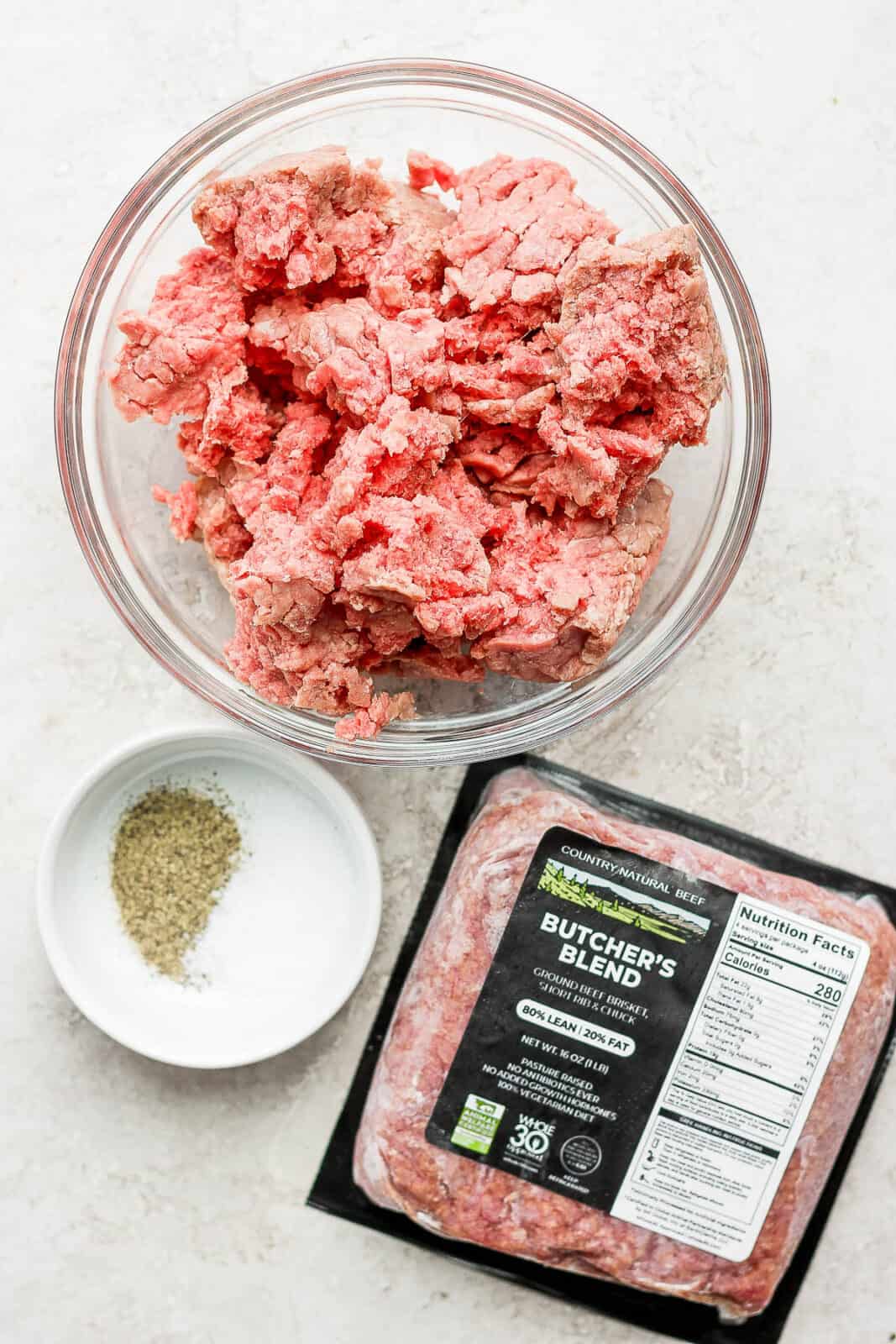 A bowl of ground beef, a bowl of salt and pepper and a package of Country Natural Beef Ground Beef.