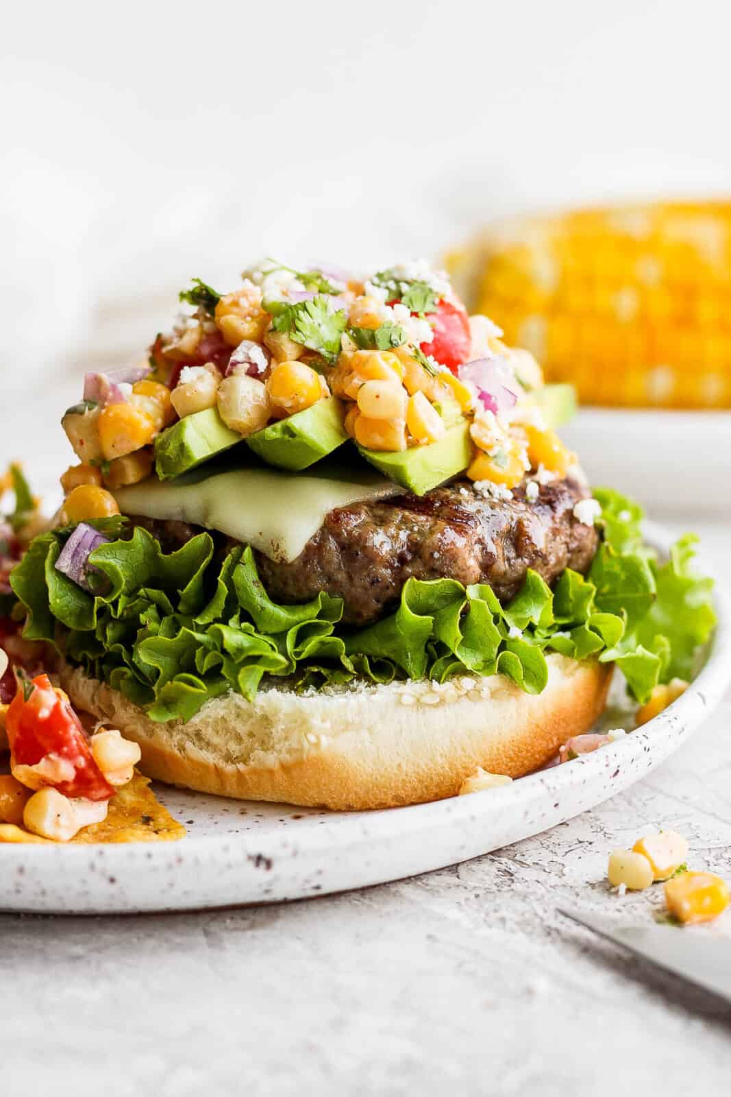 A mexican street corn burger on a plate without the top bun on yet. 