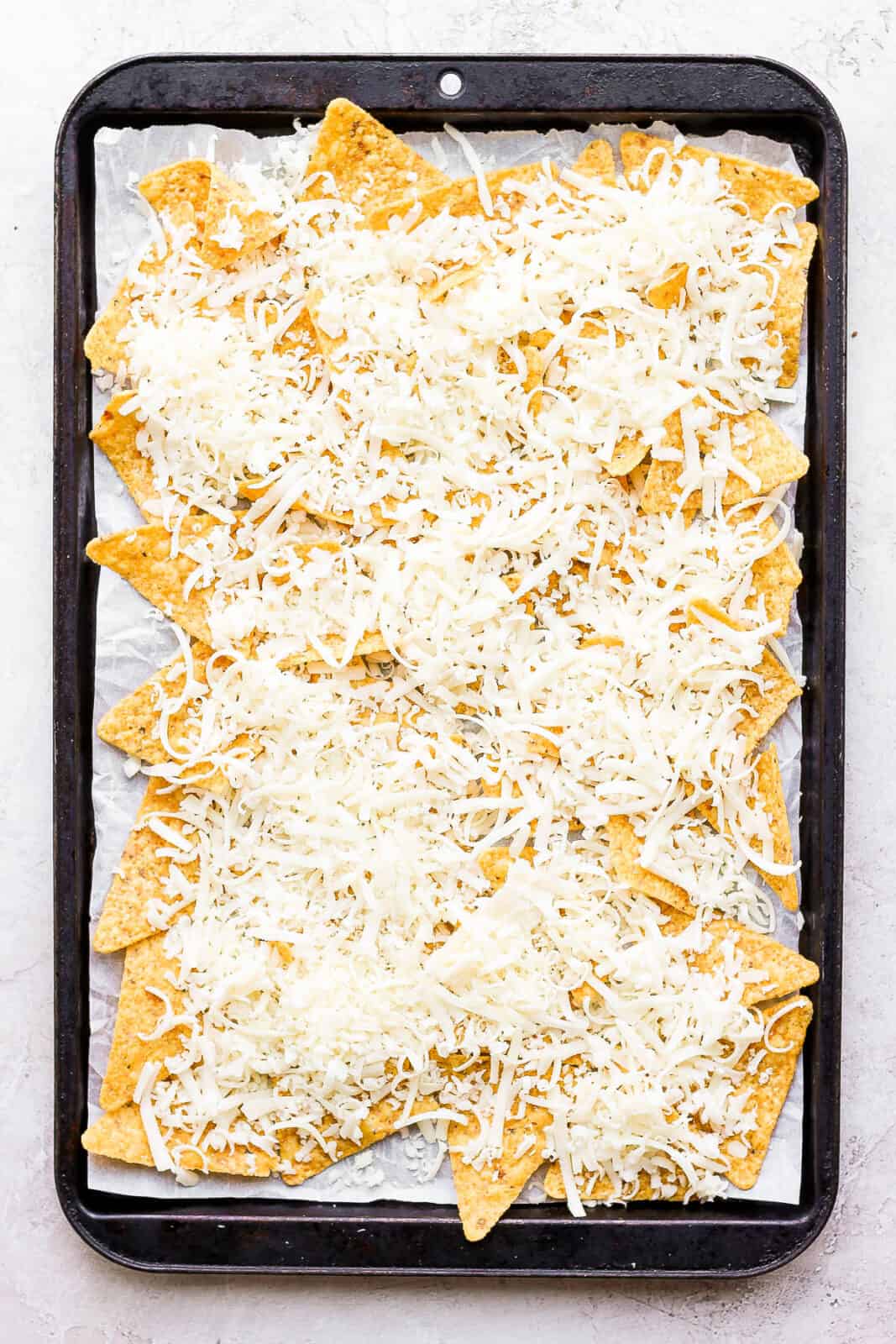 A layer of tortilla chips on a parchment-lined baking sheet with shredded cheese on top.