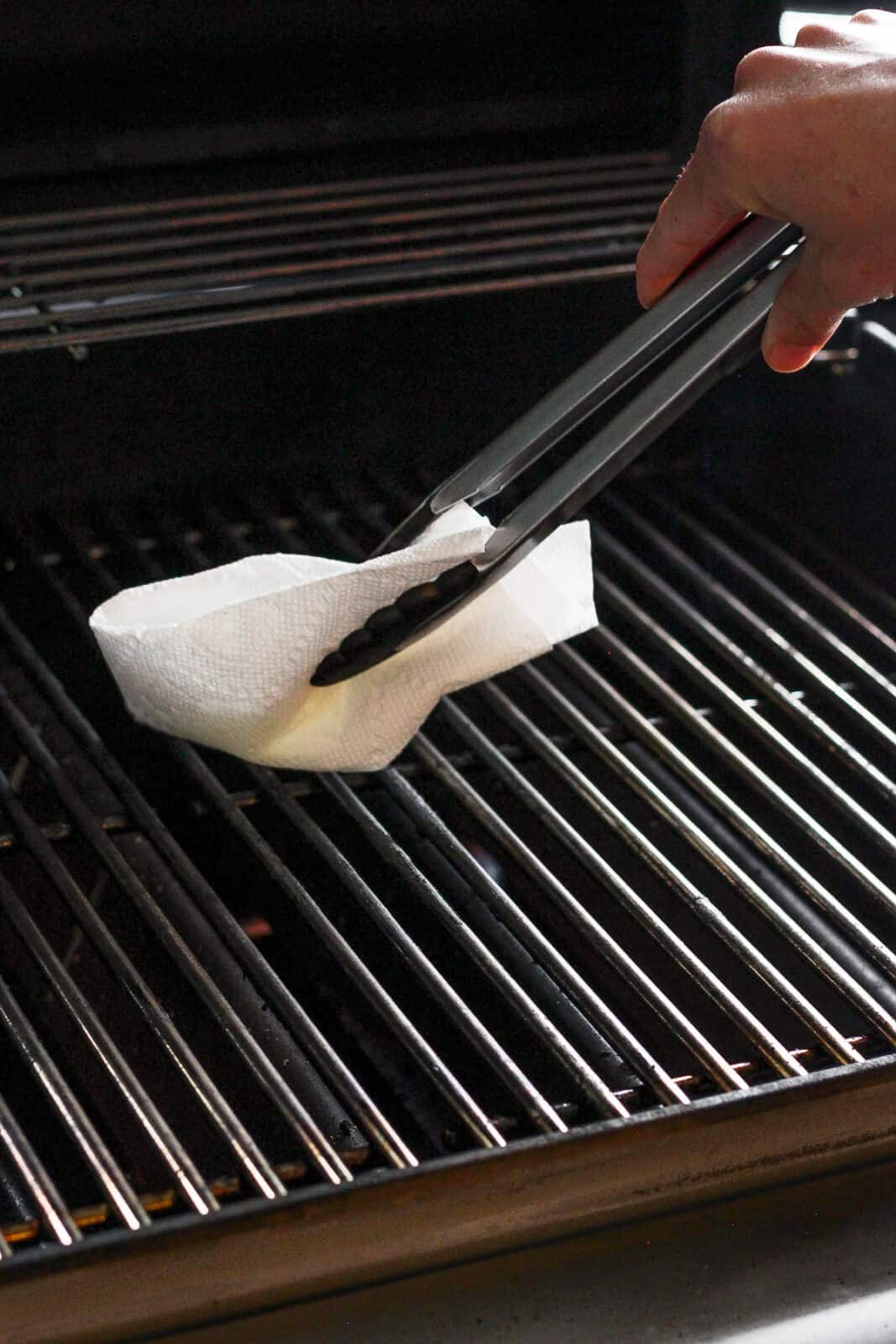 Using tongs to wipe grill grates with a paper towel coated in olive oil.