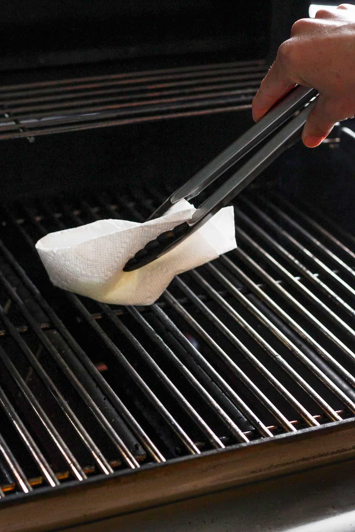 Grill grates being greased with an olive oil soaked paper towel and tongs.