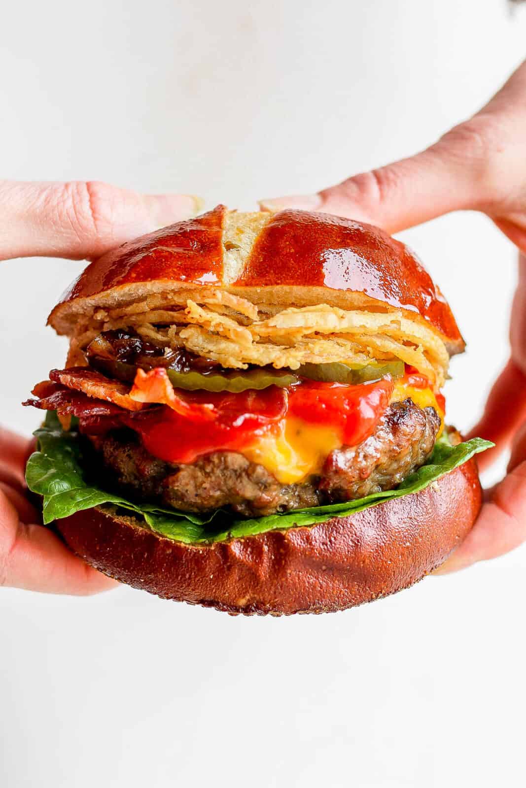 Someone holding a grilled cheeseburger on pretzel bun with fried onion strings, pickles, bacon and onion jam. 