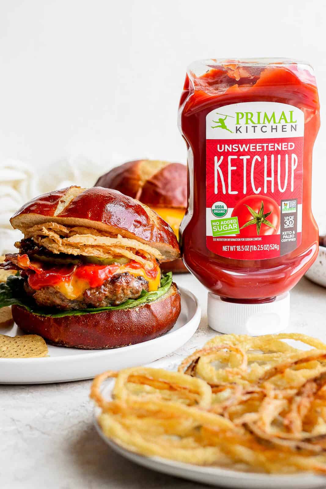 A cheeseburger on a pretzel bun next to a bottle of Primal Kitchen ketchup. 