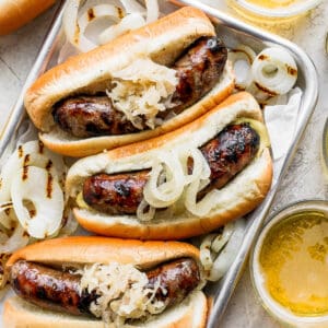 Metal tray with three beer brats in buns with onions and sauerkraut.
