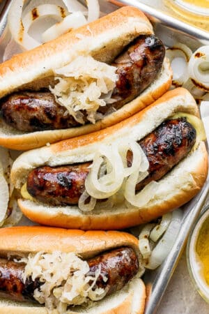 Metal tray with three beer brats in buns with onions and sauerkraut.