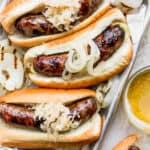 Metal tray with three beer brats in buns with onions and sauerkraut.