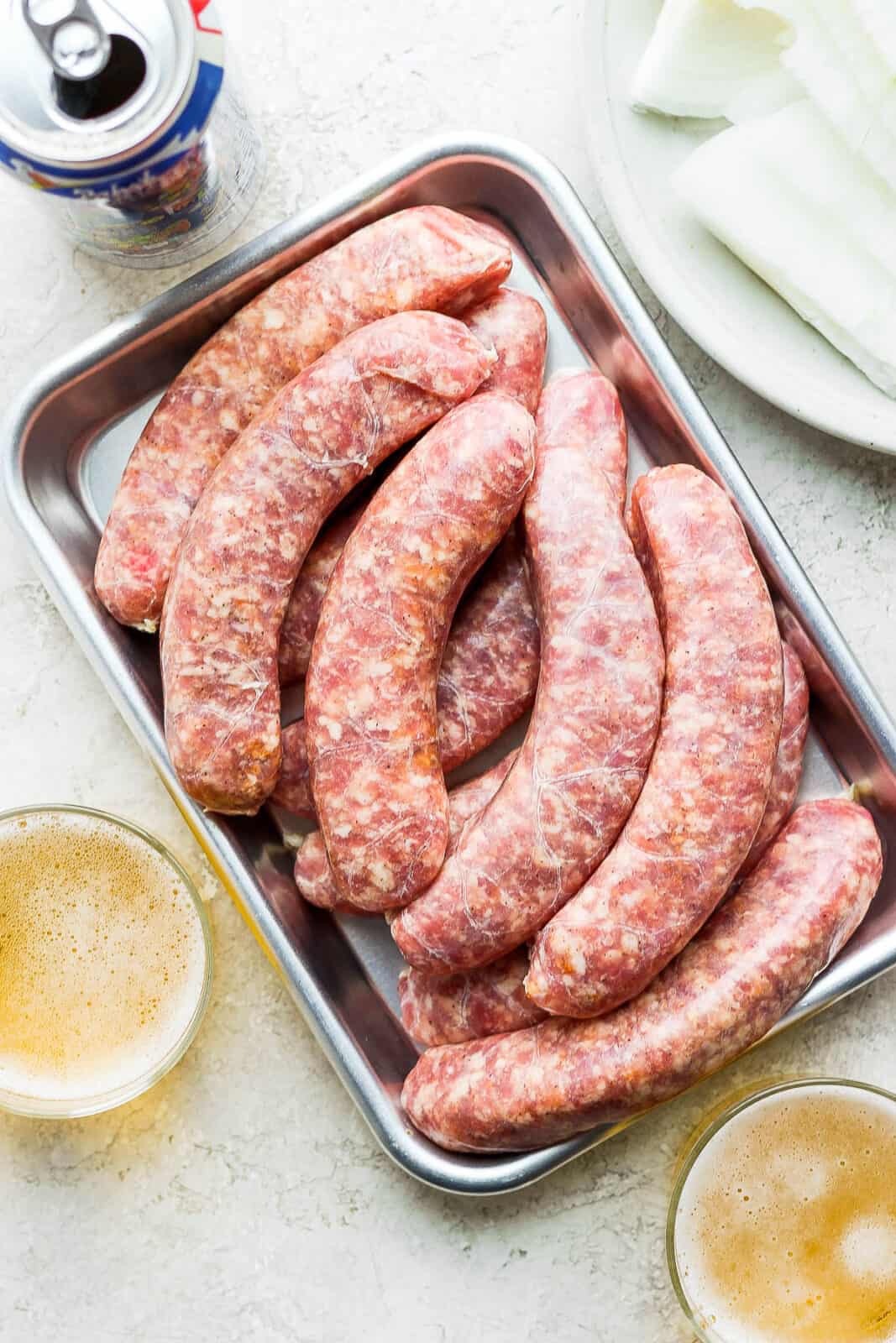 A tray of uncooked brats with some glasses of beer.