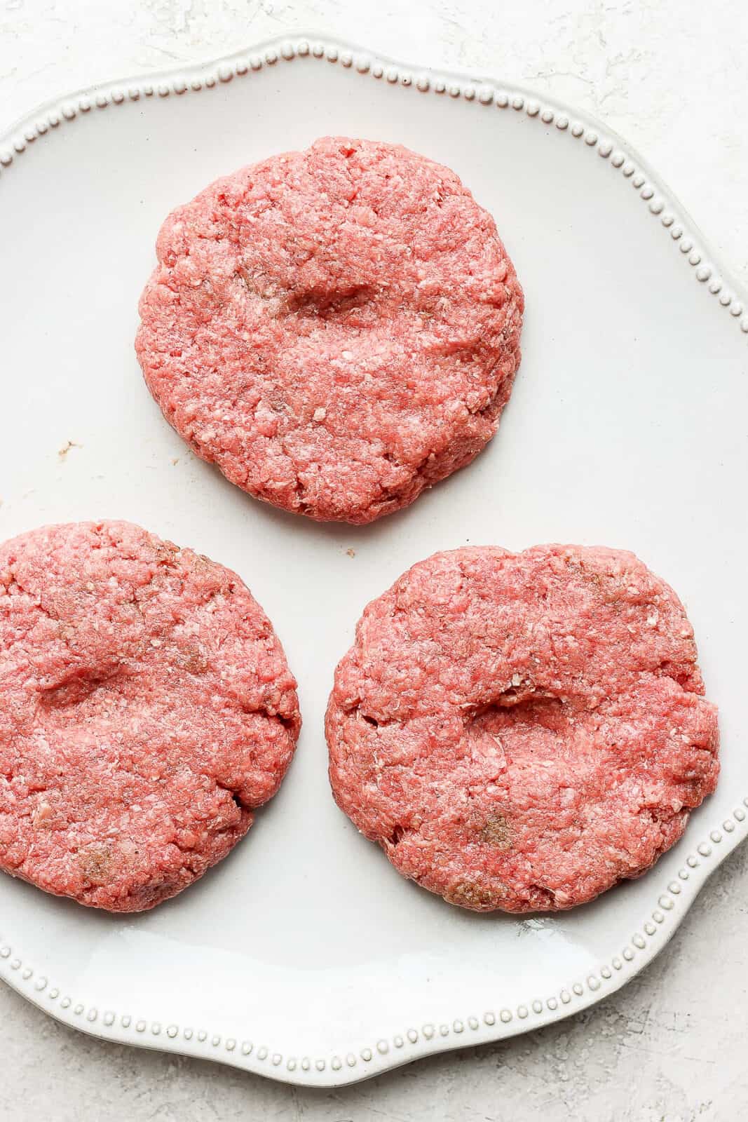 Three hamburger patties on a plate with thumbprints in the middle.