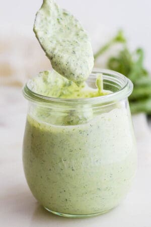 A straight on shot of a jar of green goddess dressing with a spoon scooping some out.