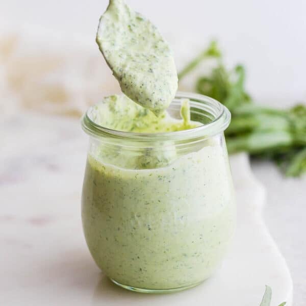 A straight on shot of a jar of green goddess dressing with a spoon scooping some out.