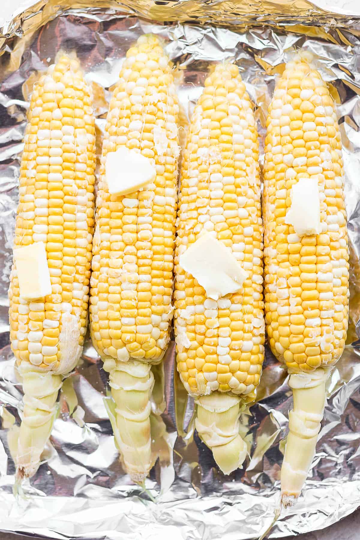 Four ears of corn on the cob laying on top of foil topped with pats of butter and kosher salt.