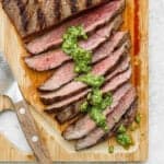 A grilled marinated flank steak on a cutting board with chimichurri sauce on it.