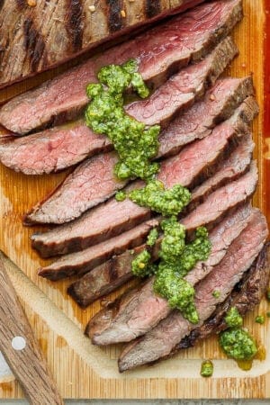 A grilled marinated flank steak on a cutting board with chimichurri sauce on it.