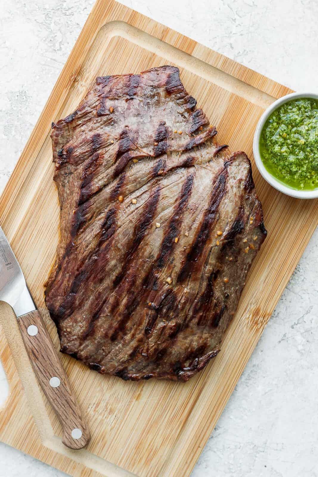 Flank steak resting on a cutting board with part of it sliced and chimichurri sauce on it. 