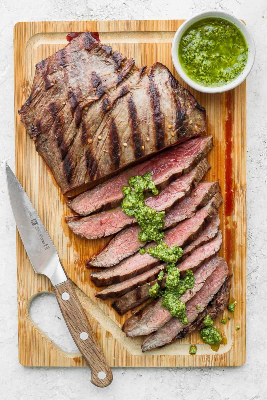 A grilled flank steak resting on a cutting board, part of it sliced with chimichurri sauce on it.