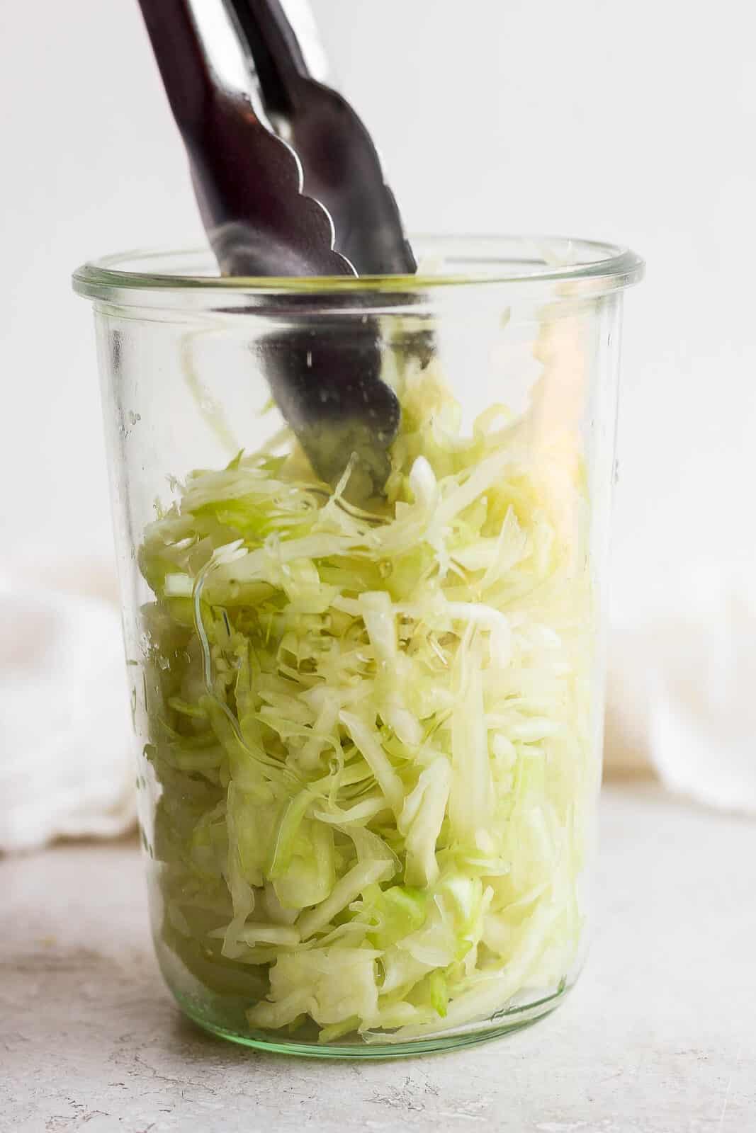 Tongs placing cabbage in a large mouth mason jar.