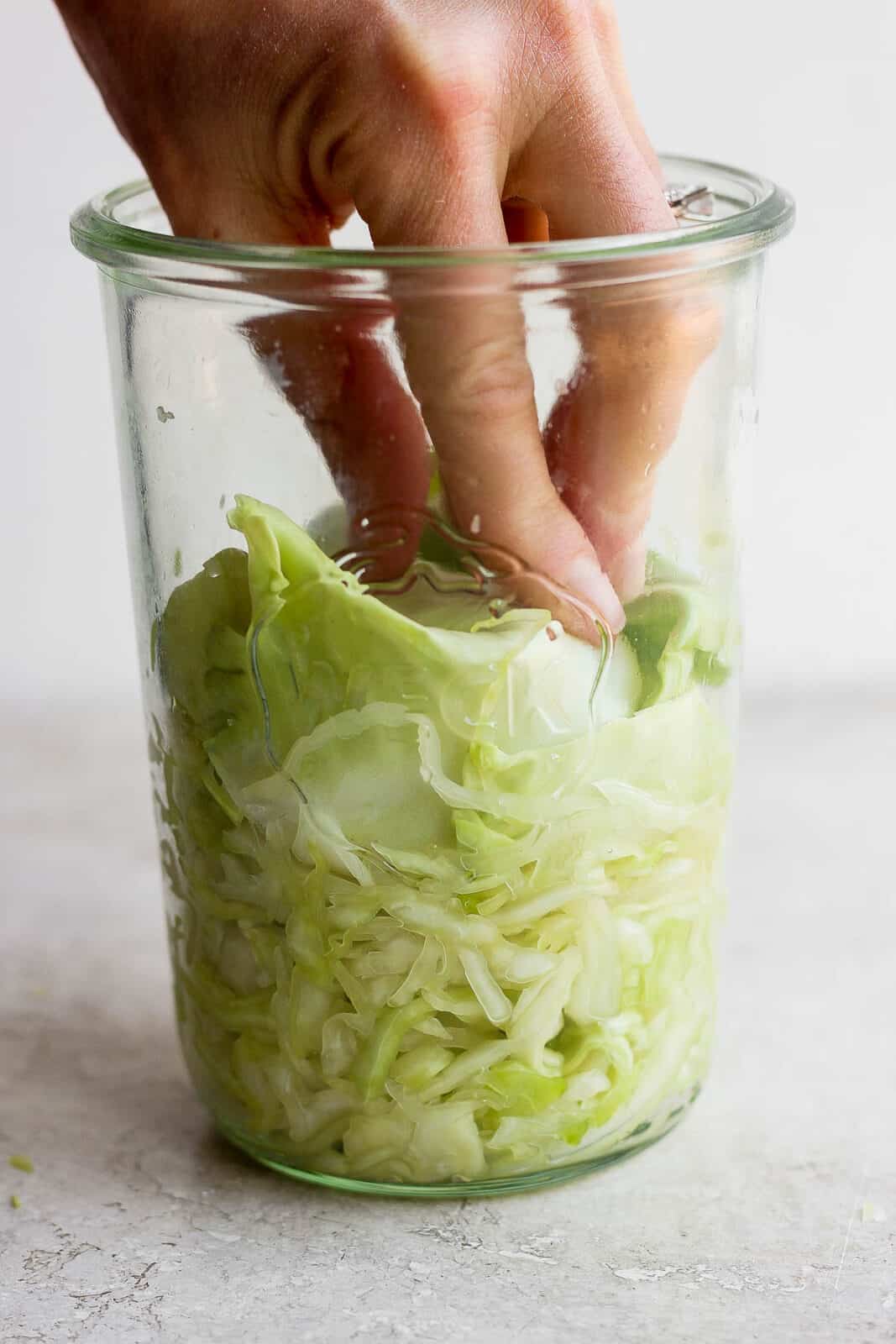 A hand pressing cabbage down in the mason jar.