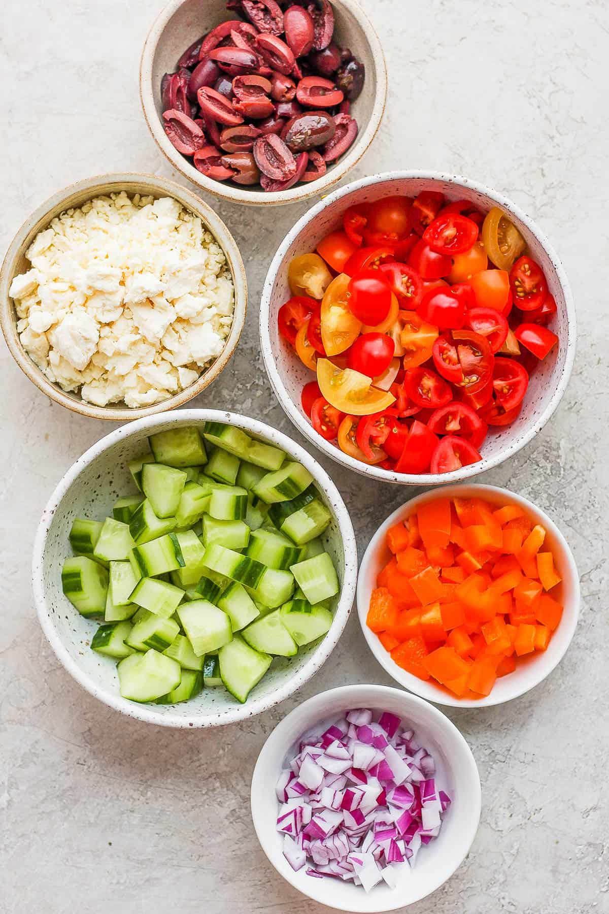 Kalamata olives, feta cheese, halved cherry tomatoes, chopped english cucumber, and chopped orange bell pepper each in their own prep bowls.