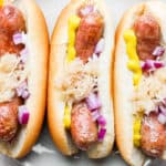 Three smoked brats in buns on a plate with various toppings,