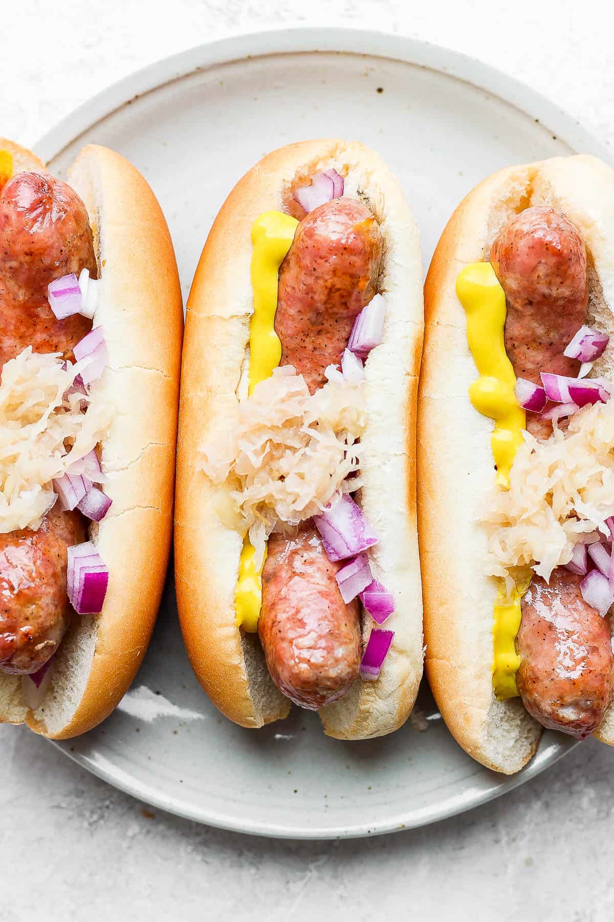 A plate of three smoked brats in buns all topped with mustard, sauerkraut and onion. 