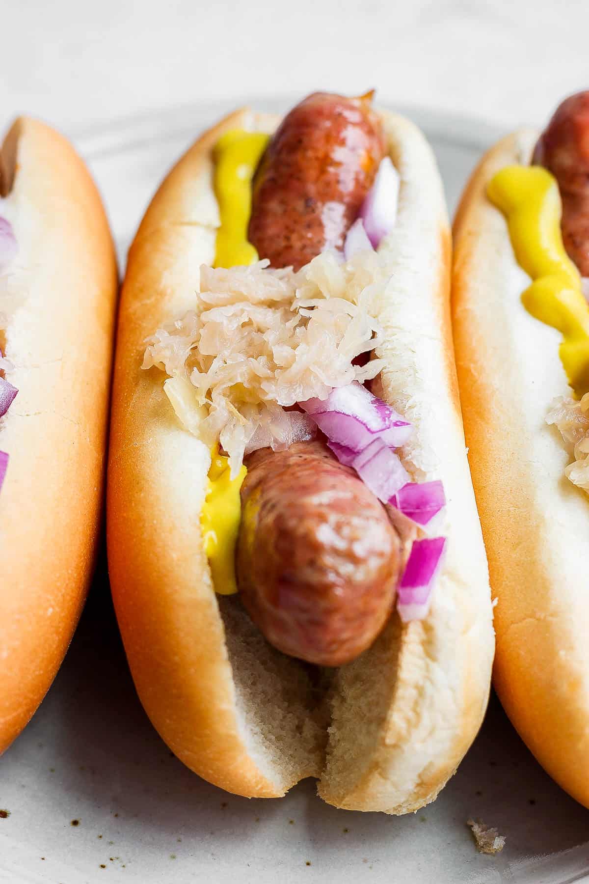 A plate of three smoked brats in buns all topped with mustard, sauerkraut and onion. 