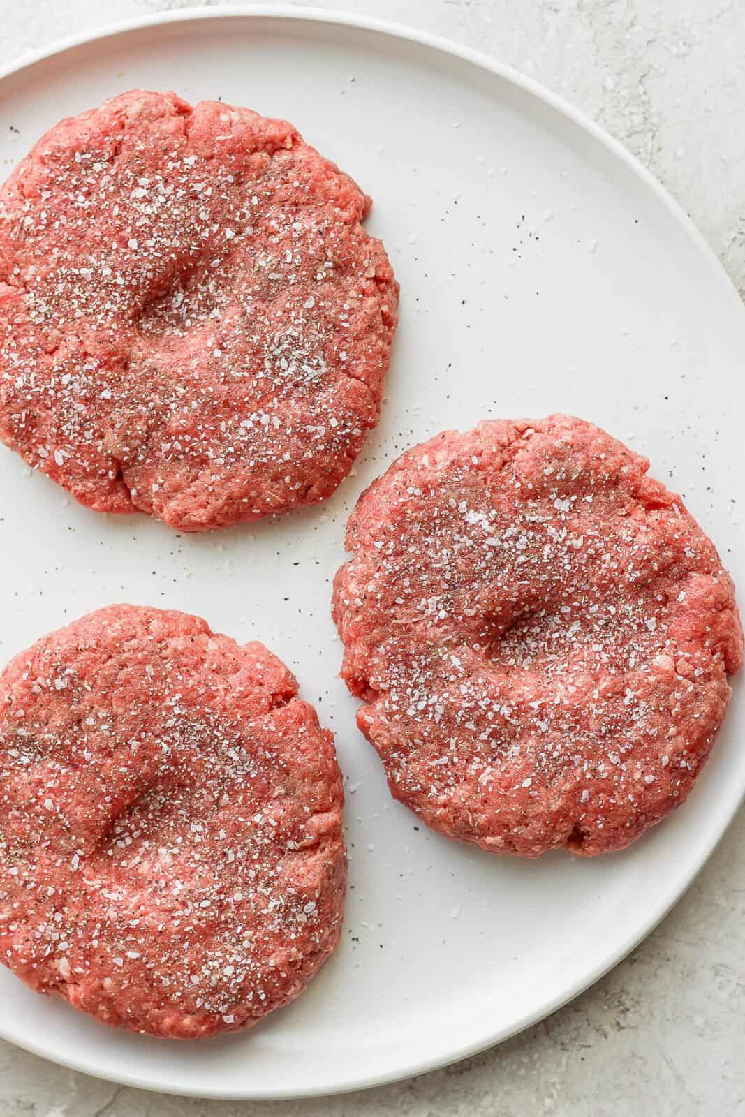 Three raw burger patties on a plate with indents in the middle and salt and pepper on top. 