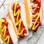 Three smoked hot dogs in buns with various toppings sitting in a wire basket.