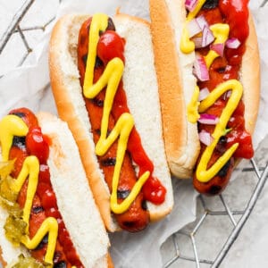 Three smoked hot dogs in buns with various toppings sitting in a wire basket.